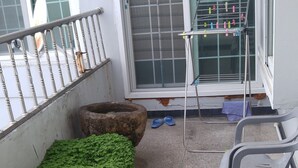 Un balcone con ringhiera in metallo, un tavolino con stendibiancheria, piante in vaso e sedie di plastica.