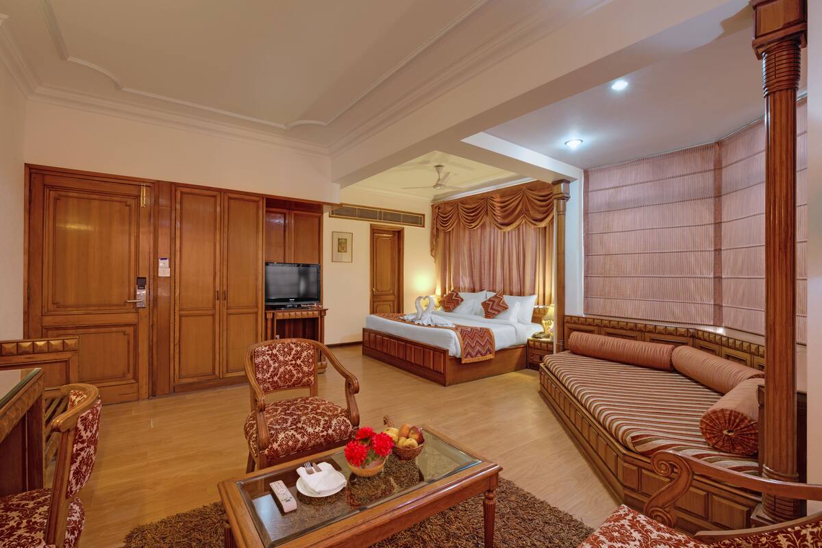 A spacious hotel room with a large bed, a sofa, a television, and a dining area with wooden furniture.