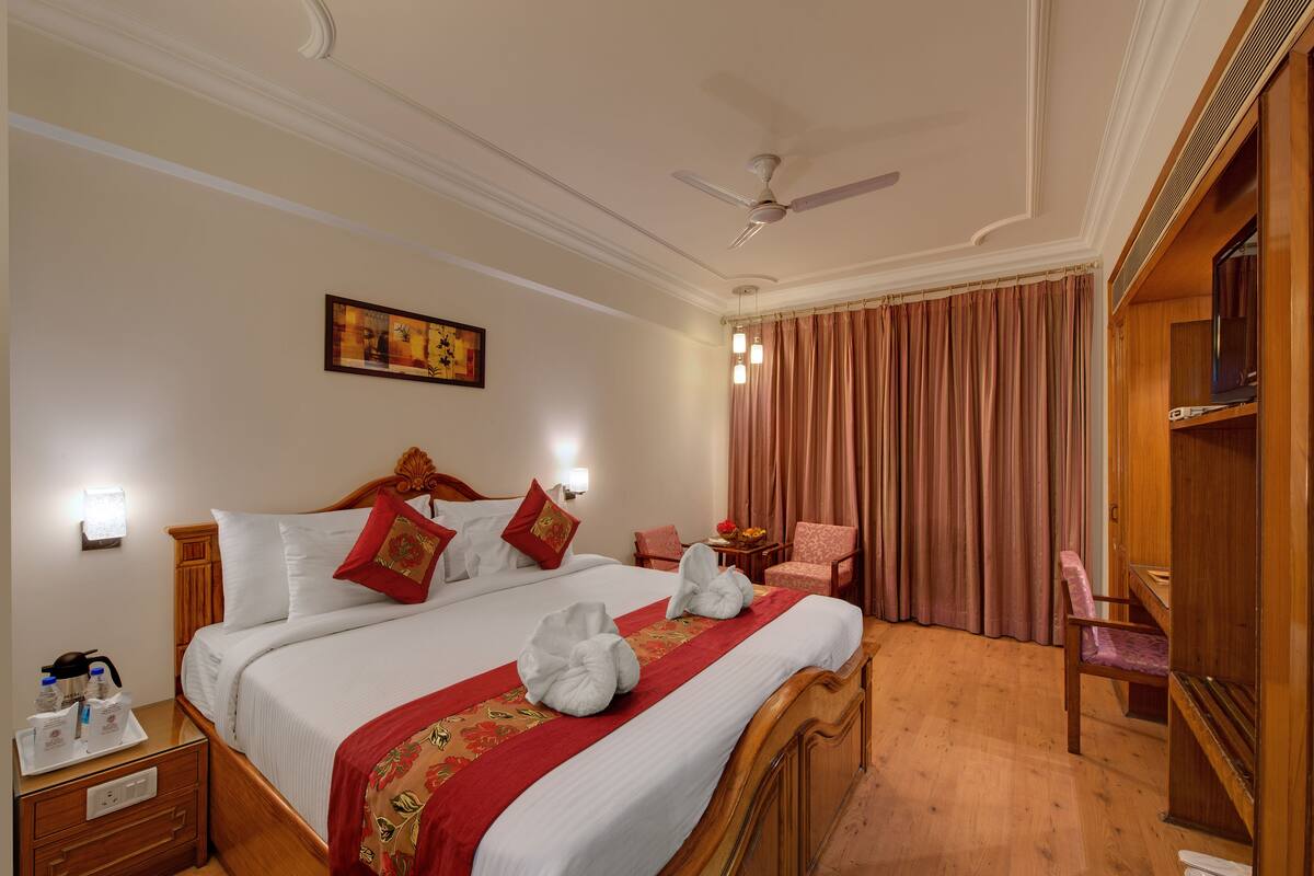 A hotel room with a large bed, a ceiling fan, a wooden wardrobe, a desk with a lamp, and a window with curtains.