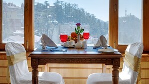 A dining table with a view of a snowy landscape through large windows, two chairs with white covers and gold sashes, and a vase with flowers and red glasses on the table. - Hotel Baljees Regency (Shimla)