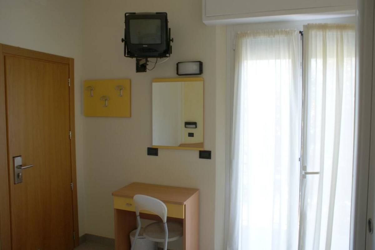 A room with a wooden door, a desk with a chair, a television mounted on the wall, a mirror, and a window with curtains.