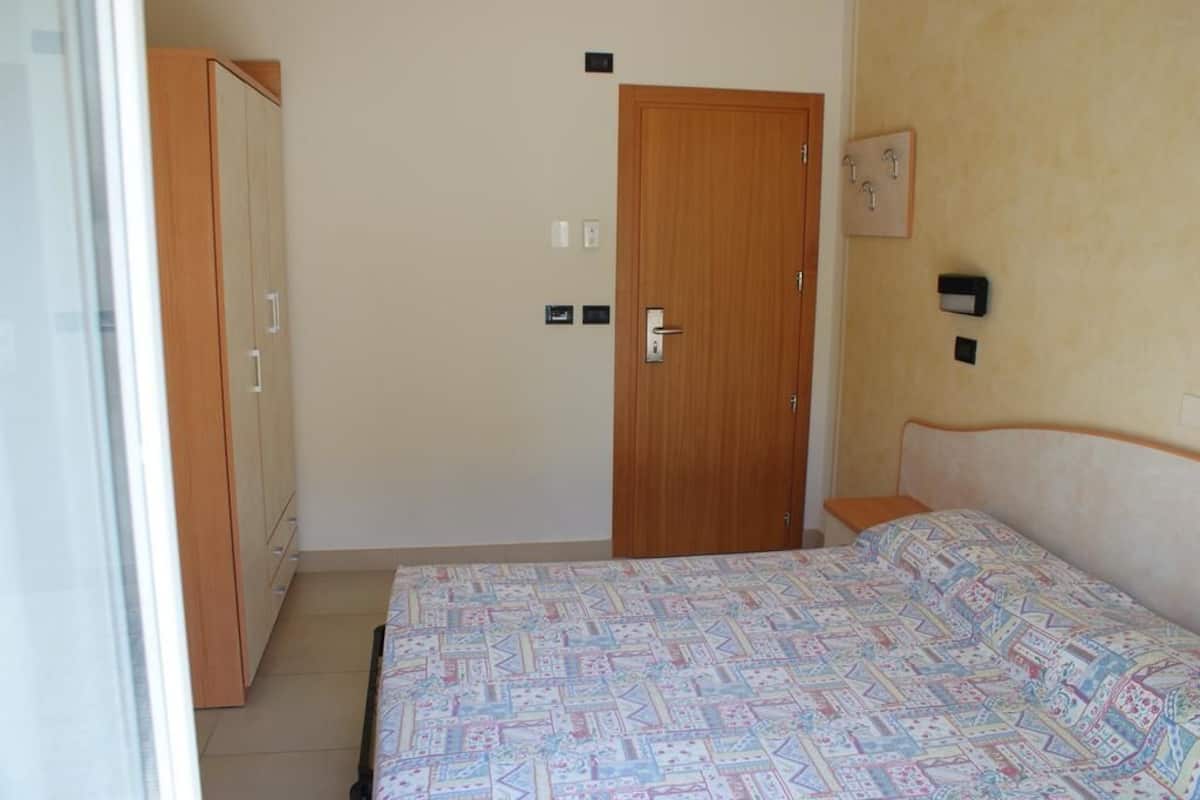 A bedroom with a bed, a wardrobe, a door, and a wall-mounted light switch.
