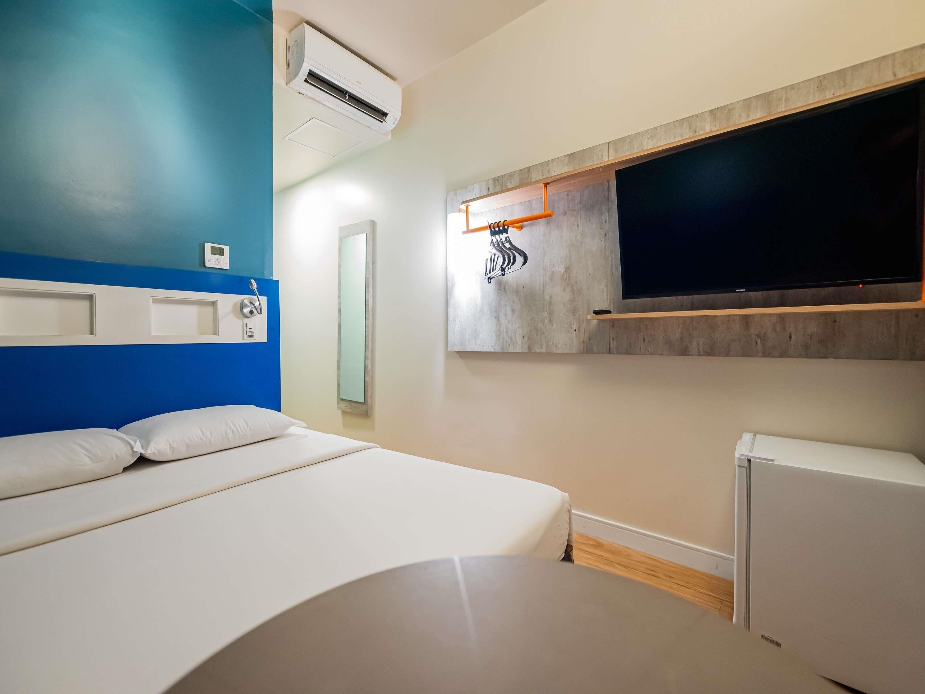 A hotel room with a bed, a wall-mounted TV, an air conditioning unit, and a small refrigerator.