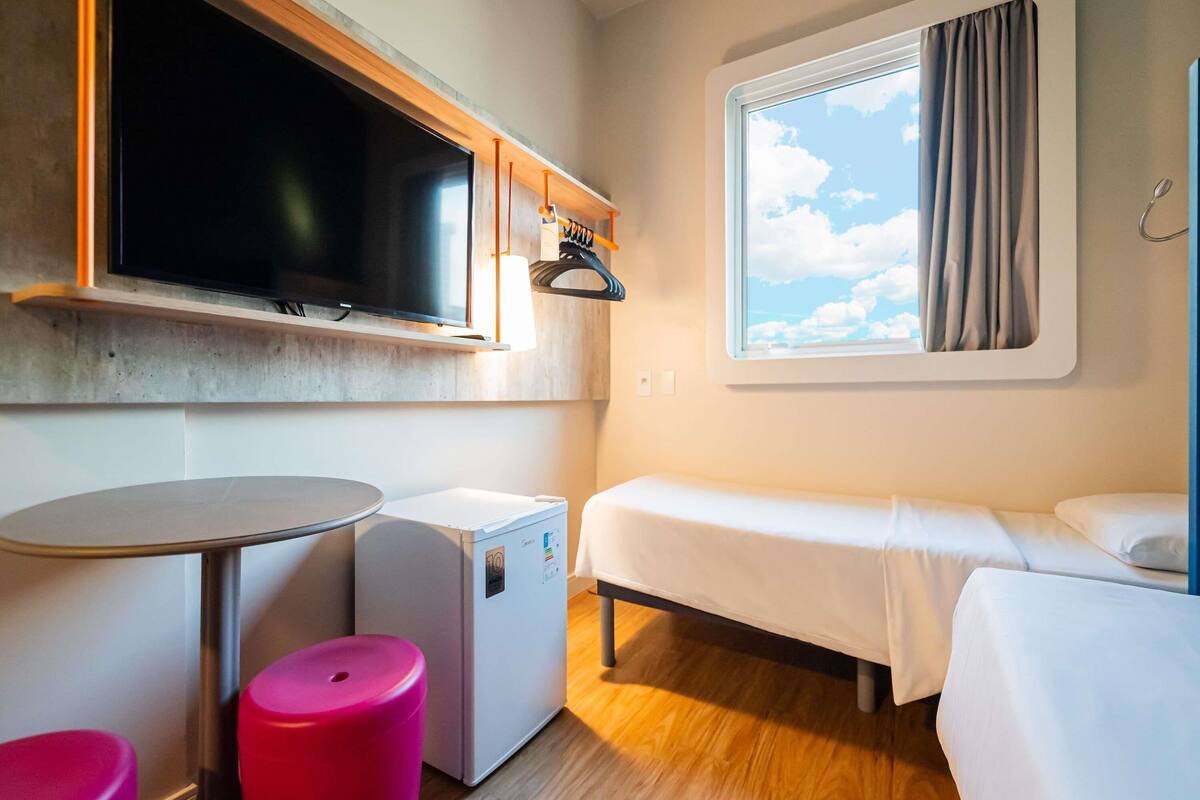 Een compacte hotelkamer met een bed, een klein tafeltje, een koelkast, een aan de muur bevestigde televisie en een raam met gordijnen.