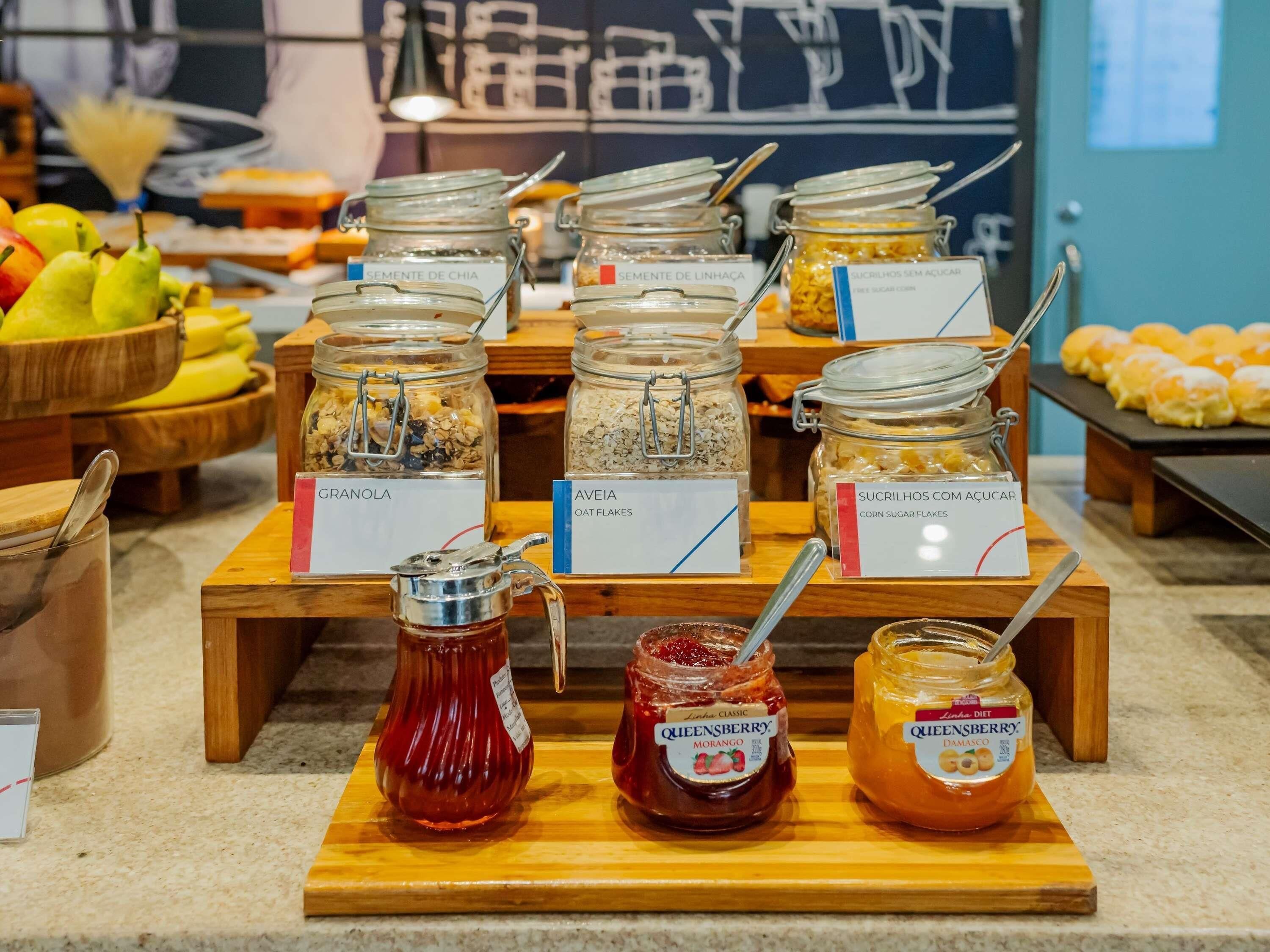 Buffet de café da manhã com alimentos identificados, incluindo granola, flocos de aveia, flocos de milho açucarados, sementes de chia e linhaça.