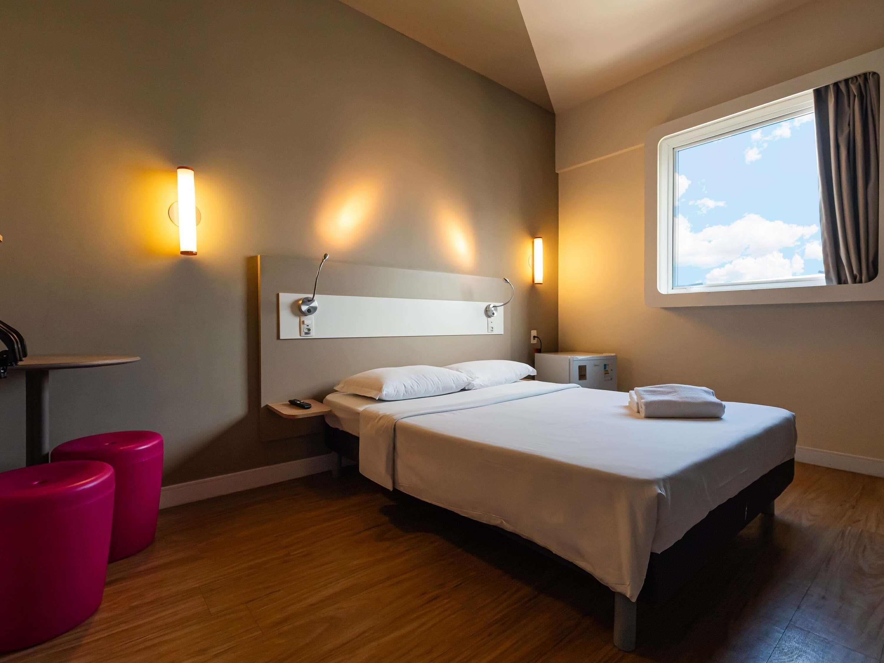 A hotel room with a bed, bedside table, lamp, and a window with curtains.