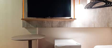 Quarto com uma cama, uma mesa pequena, dois banquinhos vermelhos, um armário branco e uma televisão na parede.