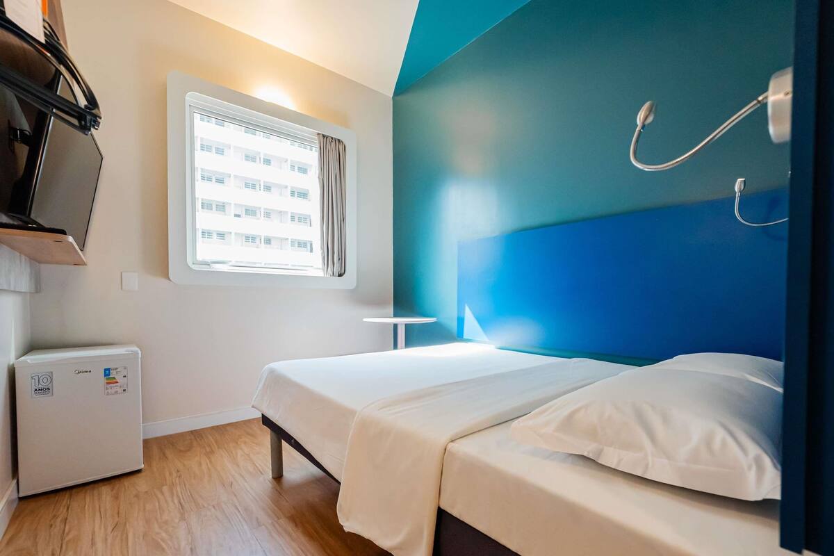Een kleine, moderne kamer met een bed, een kleine koelkast, een raam met zonwering en een blauwe accentmuur.