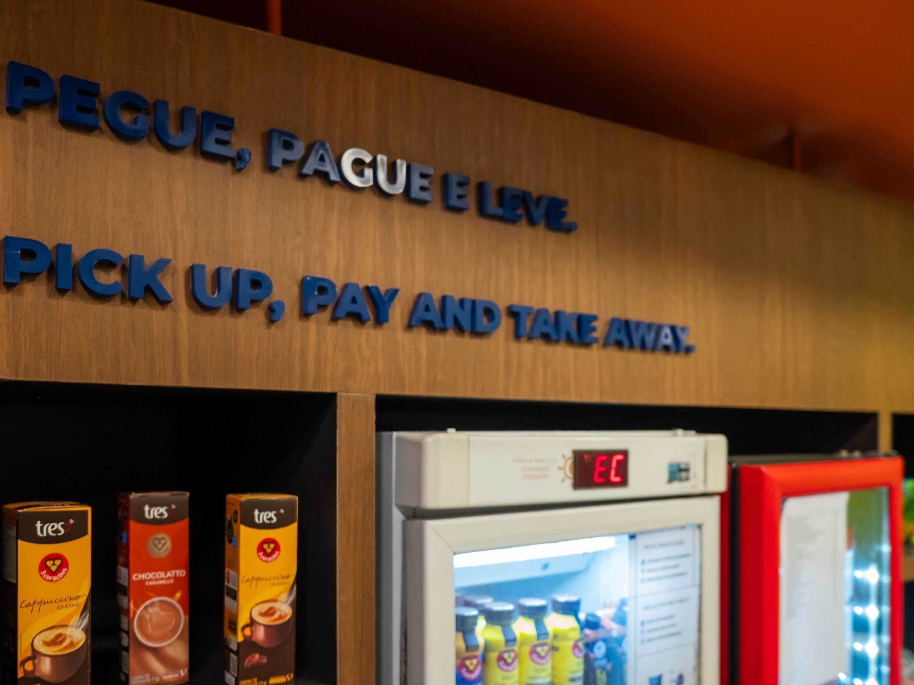 A sign with 'Pique, Pague e Leve. Pick up, Pay and Take Away.' and a refrigerator with beverages.