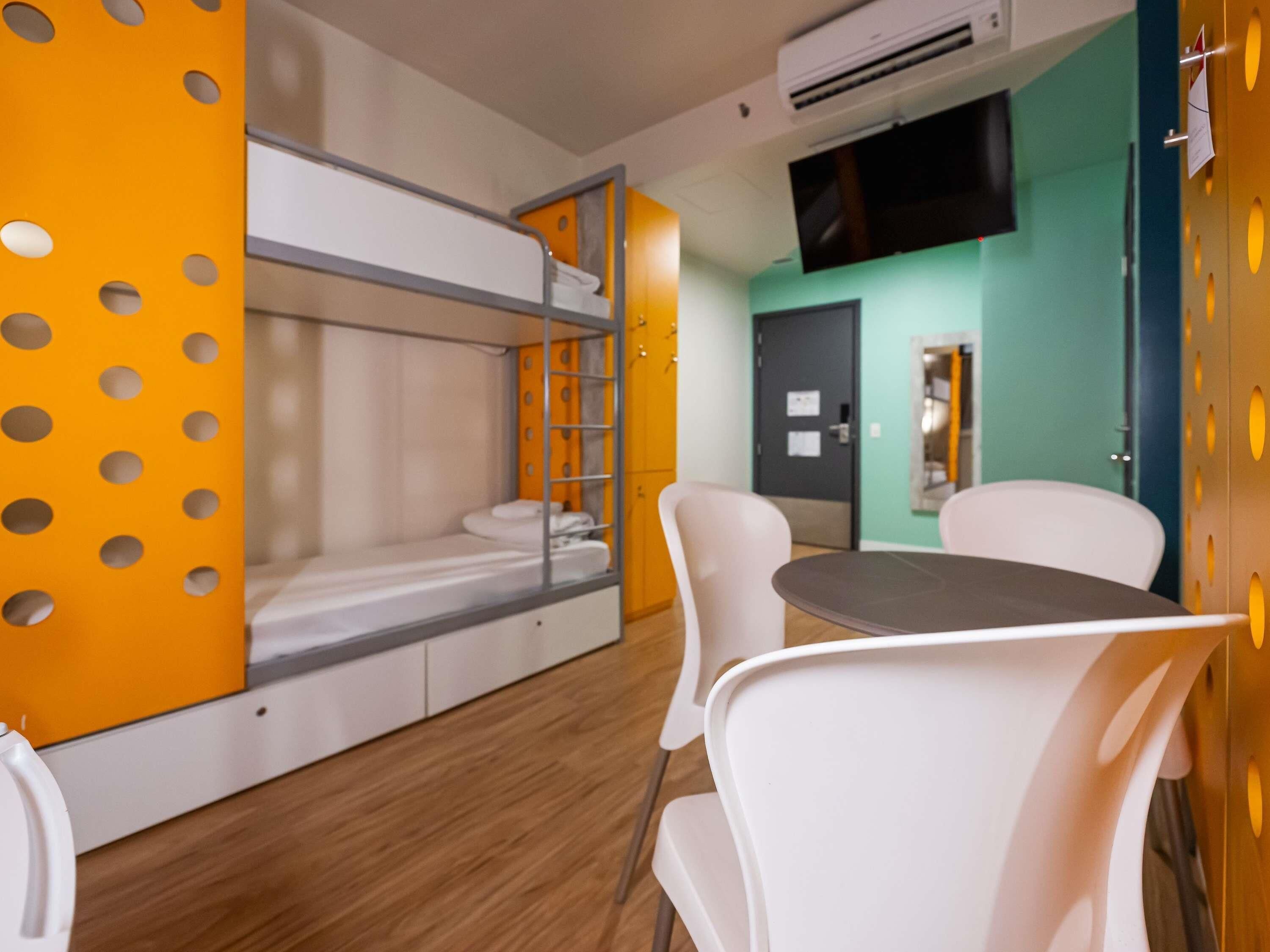 A bunk bed with a white mattress, a small table, and white chairs in a room with wooden flooring and a green accent wall.