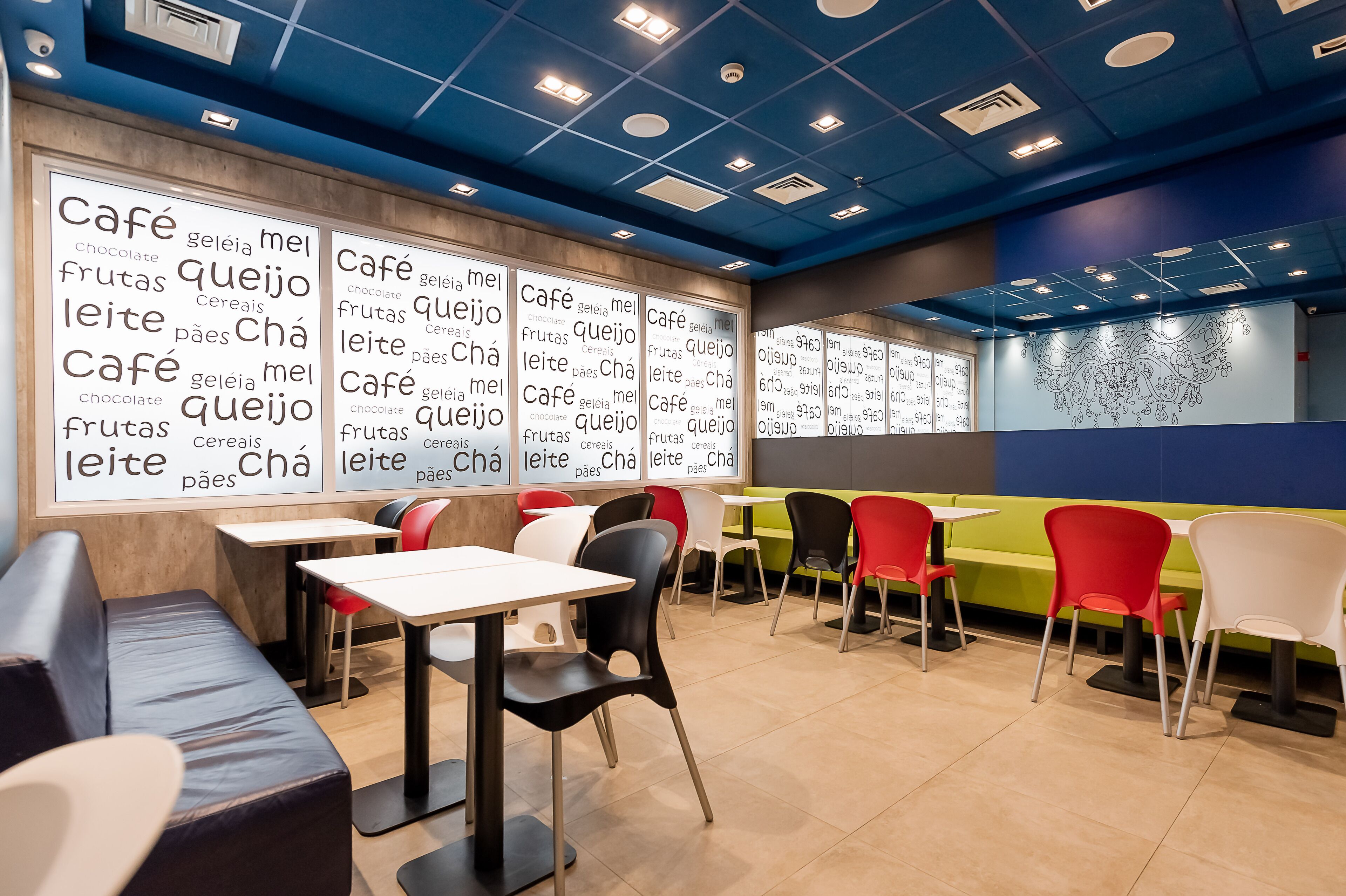 A café with tables and chairs, a wall with words in a foreign language, and a blue ceiling with lights.