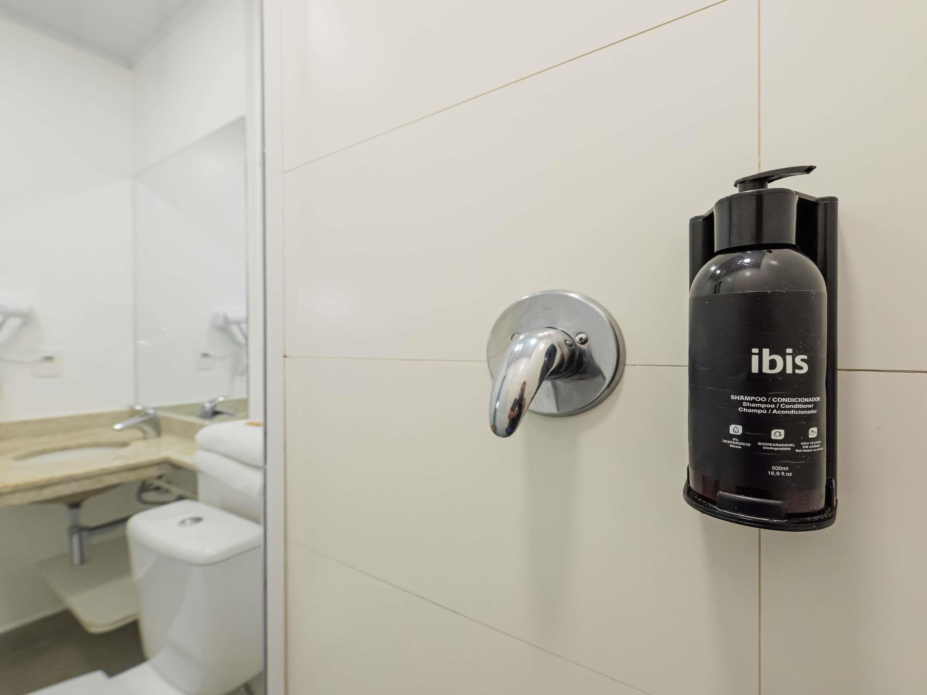 A bathroom with a white tiled wall, a chrome faucet, and a black bottle of Ibis shampoo.