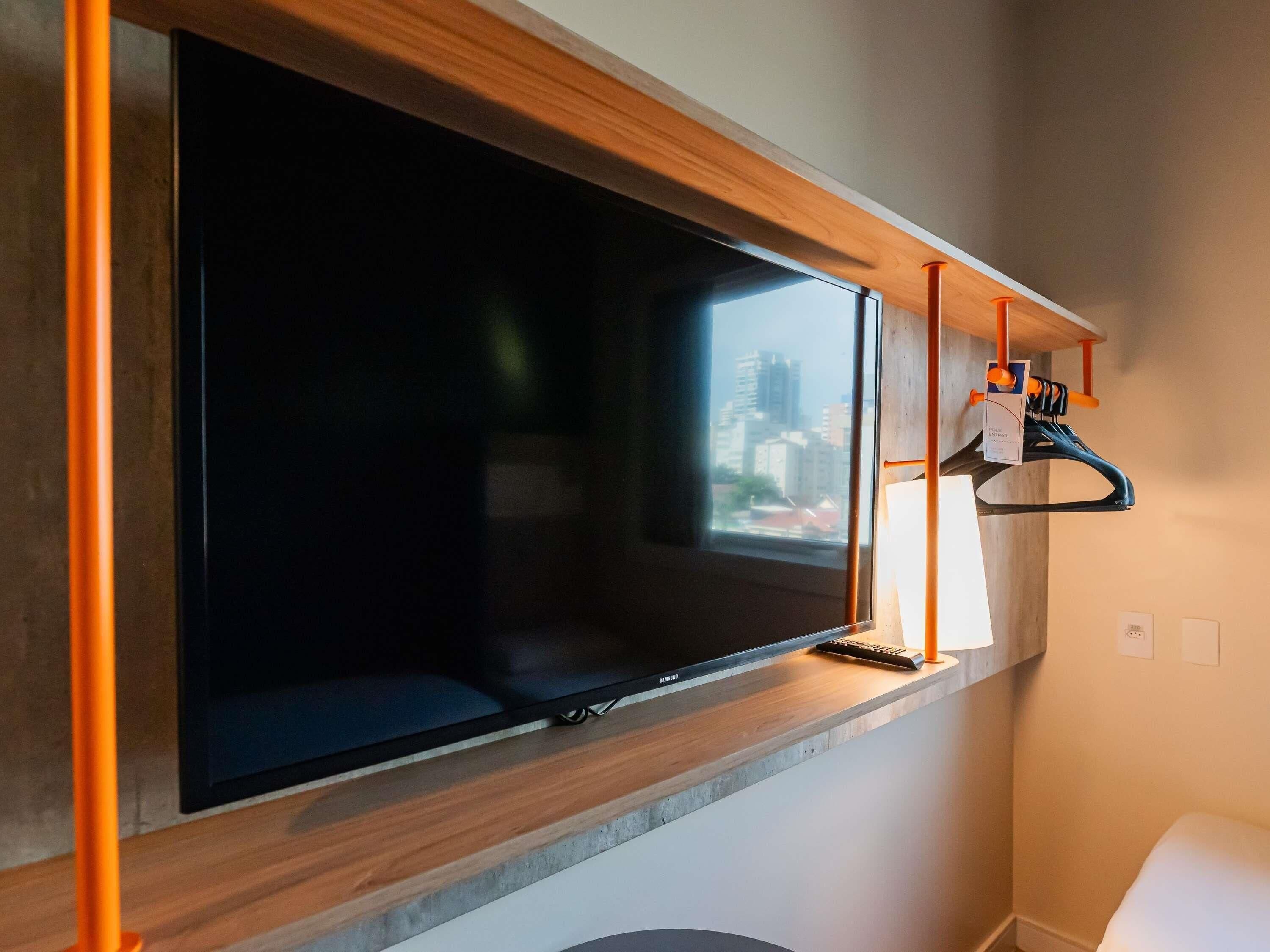 A modern room with a large flat-screen TV mounted on a wooden shelf, a black remote control, and a black hanger on the wall.