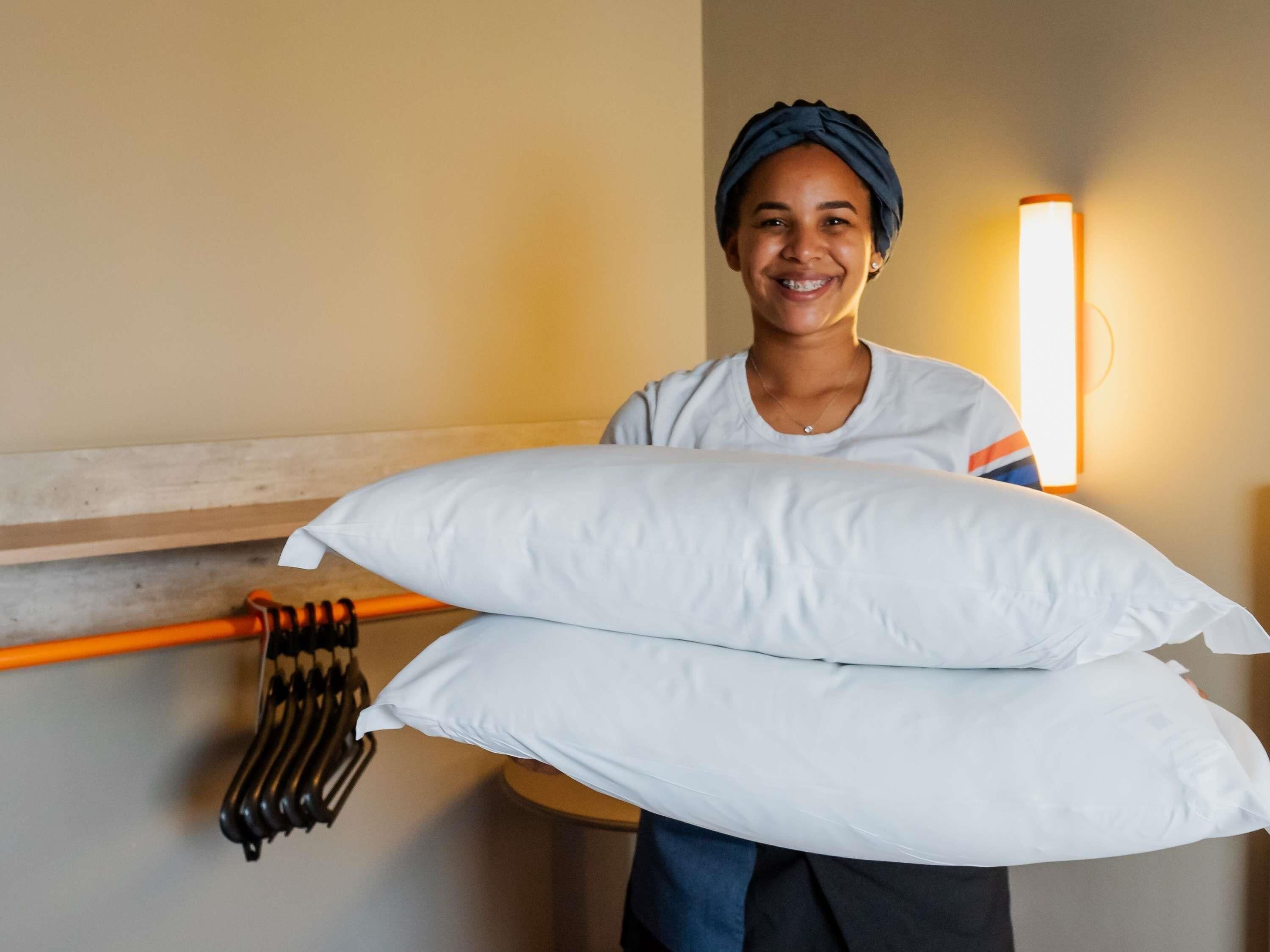 A person holding two white pillows, a wooden headboard, and a wall-mounted light fixture.