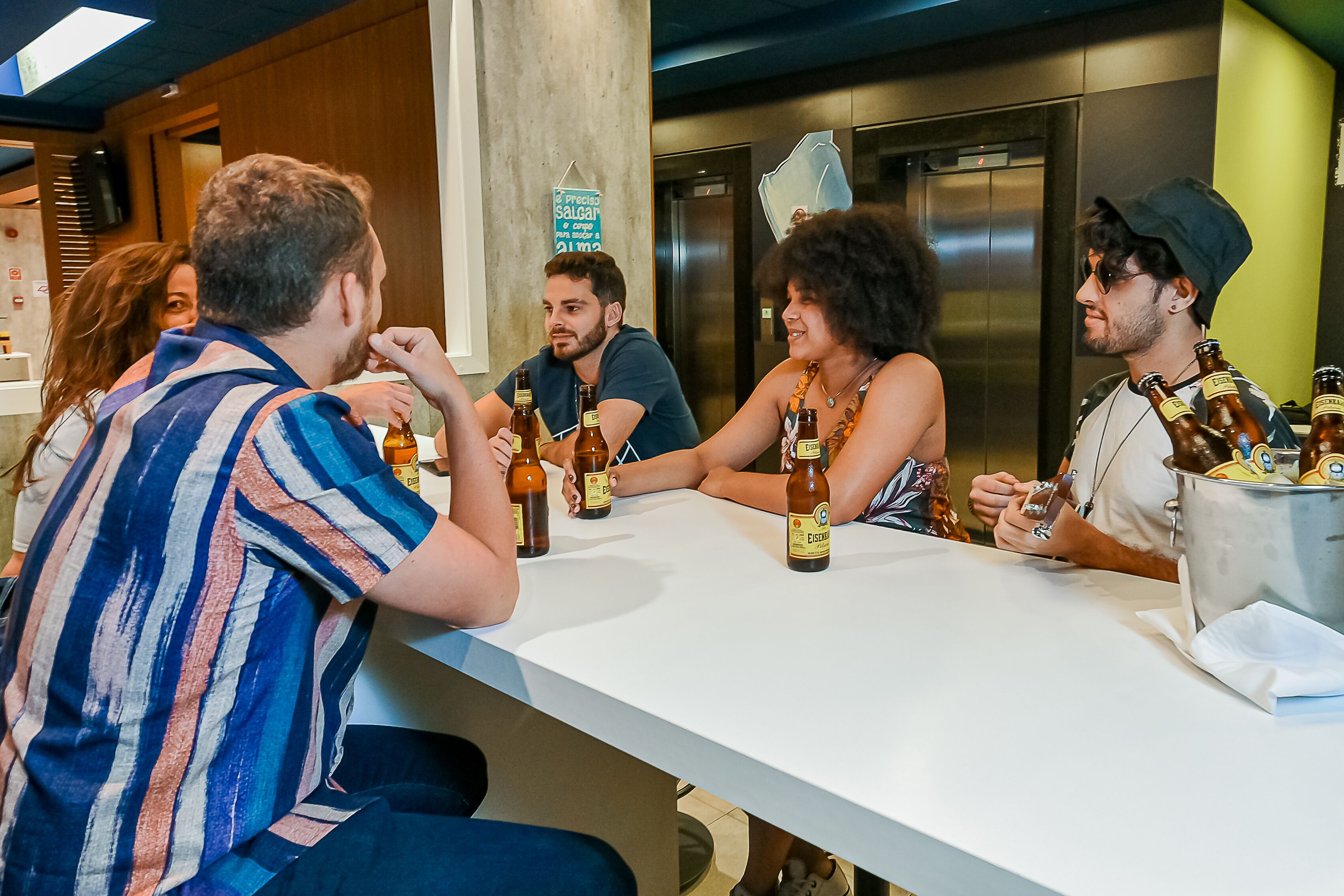 A group of people sitting at a table with bottles of beer, in a room with a white table and a sign on the wall.