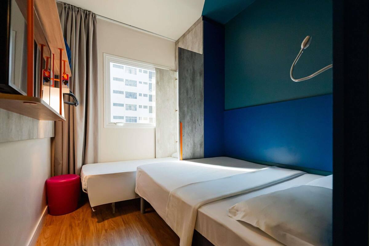 Een hotelkamer met twee bedden, een raam, een rode kruk en een hangende douchekop.