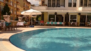 Uma piscina com borda azulejada, rodeada por área de estar ao ar livre com guarda-sóis e mesas, e um edifício com varandas ao fundo.