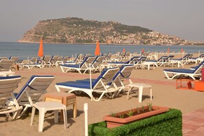 A beach with rows of sun loungers and umbrellas, a clear view of the sea, and a hillside in the background.