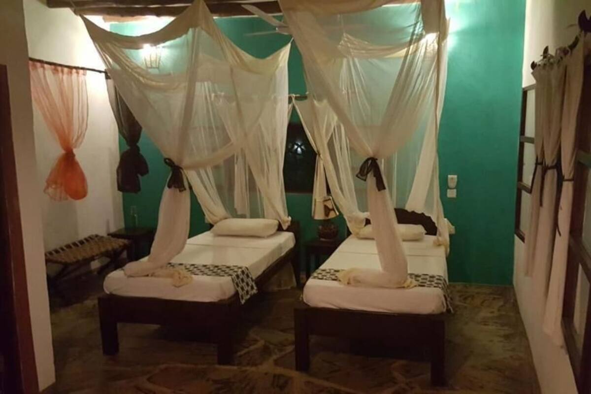 Habitación con dos camas, cada una con dosel. Hay dos mesitas de noche y un banco. La pared es verde y la alfombra tiene un estampado.