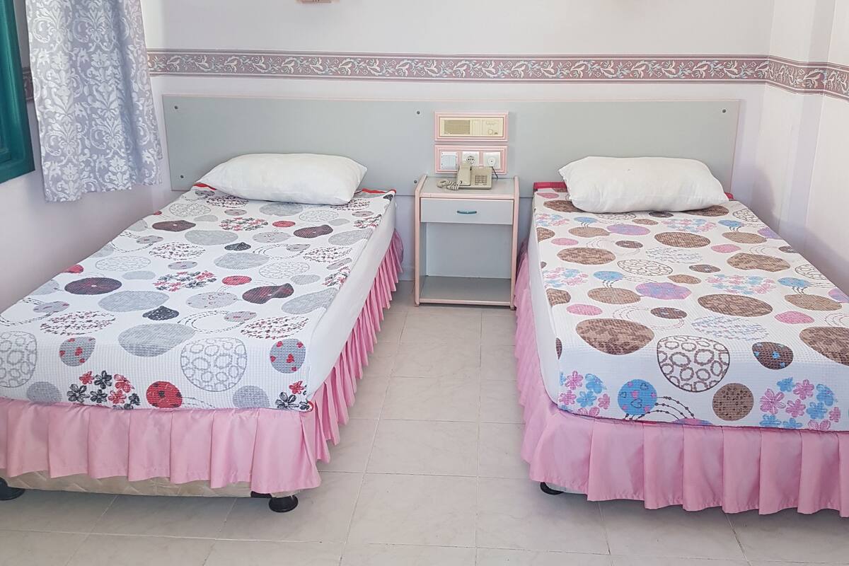 A hotel room with two single beds, each with a patterned bedspread and pink trim. There is a bedside table with a telephone and a small lamp. The room has a simple, clean design with a plain wall and a small picture frame.
