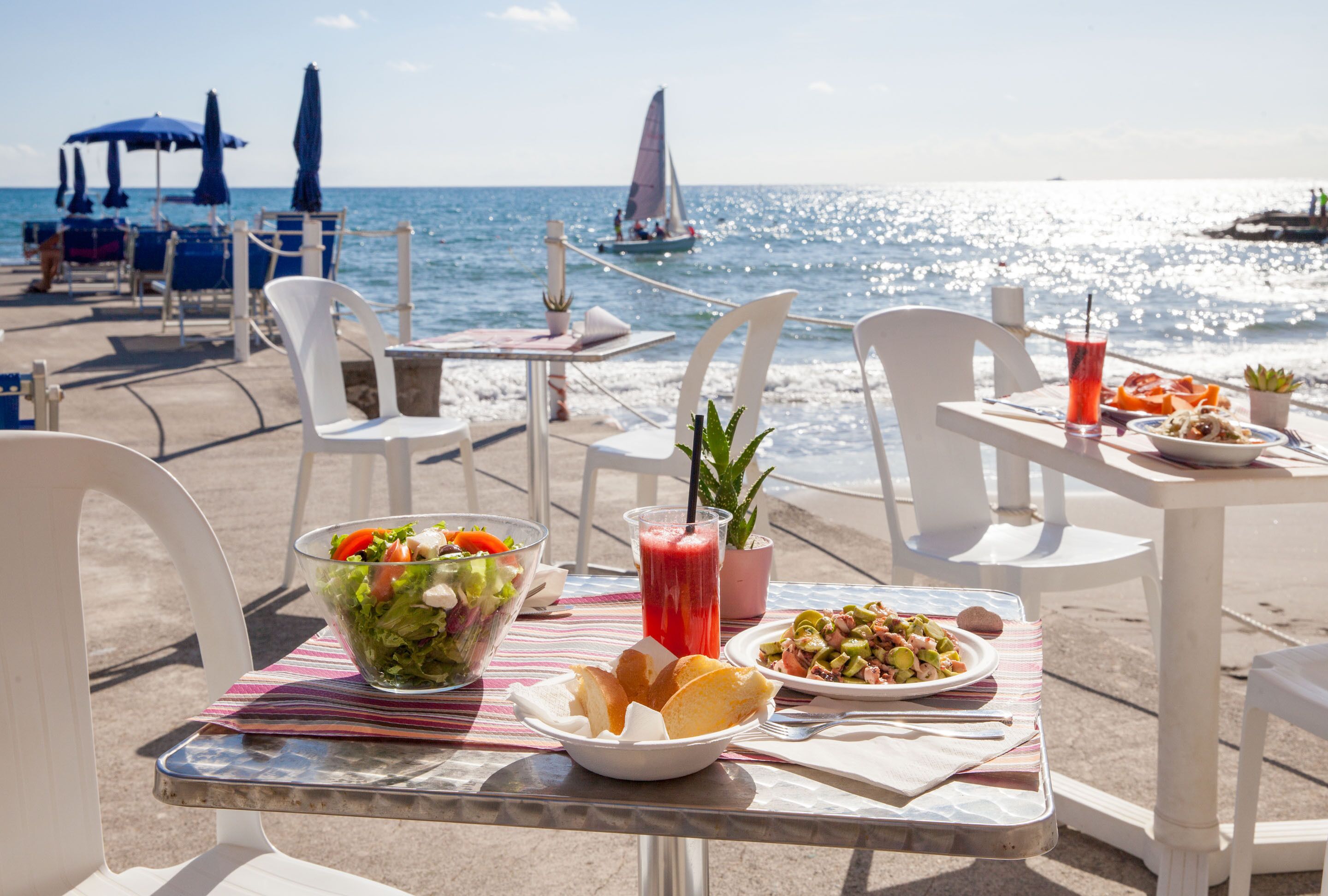 A table with a salad, a drink, and bread, set on a beach with chairs and umbrellas.