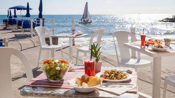 A table with a salad, a drink, and bread, set on a beach with chairs and umbrellas.