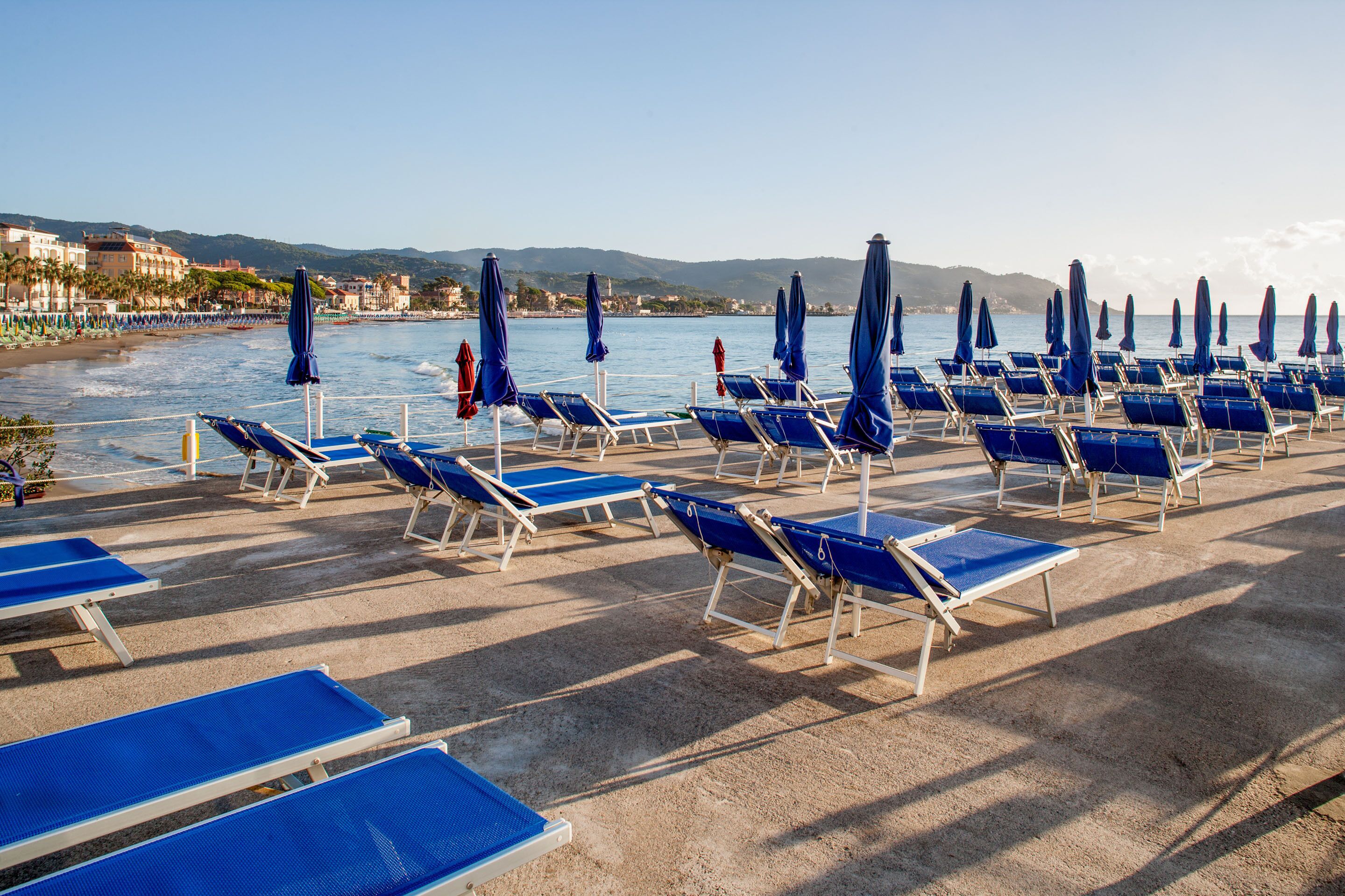 A beachside lounge area with rows of blue lounge chairs and umbrellas, a clear view of the sea, and a distant mountain range.