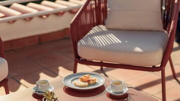 Um terraço com uma cadeira de vime, uma mesa com uma xícara de café e um prato de comida, e vista para o mar.