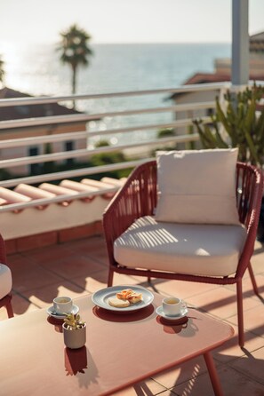 Una terraza con una silla de mimbre, una mesa con una taza de café y un plato de comida, y vistas al mar.