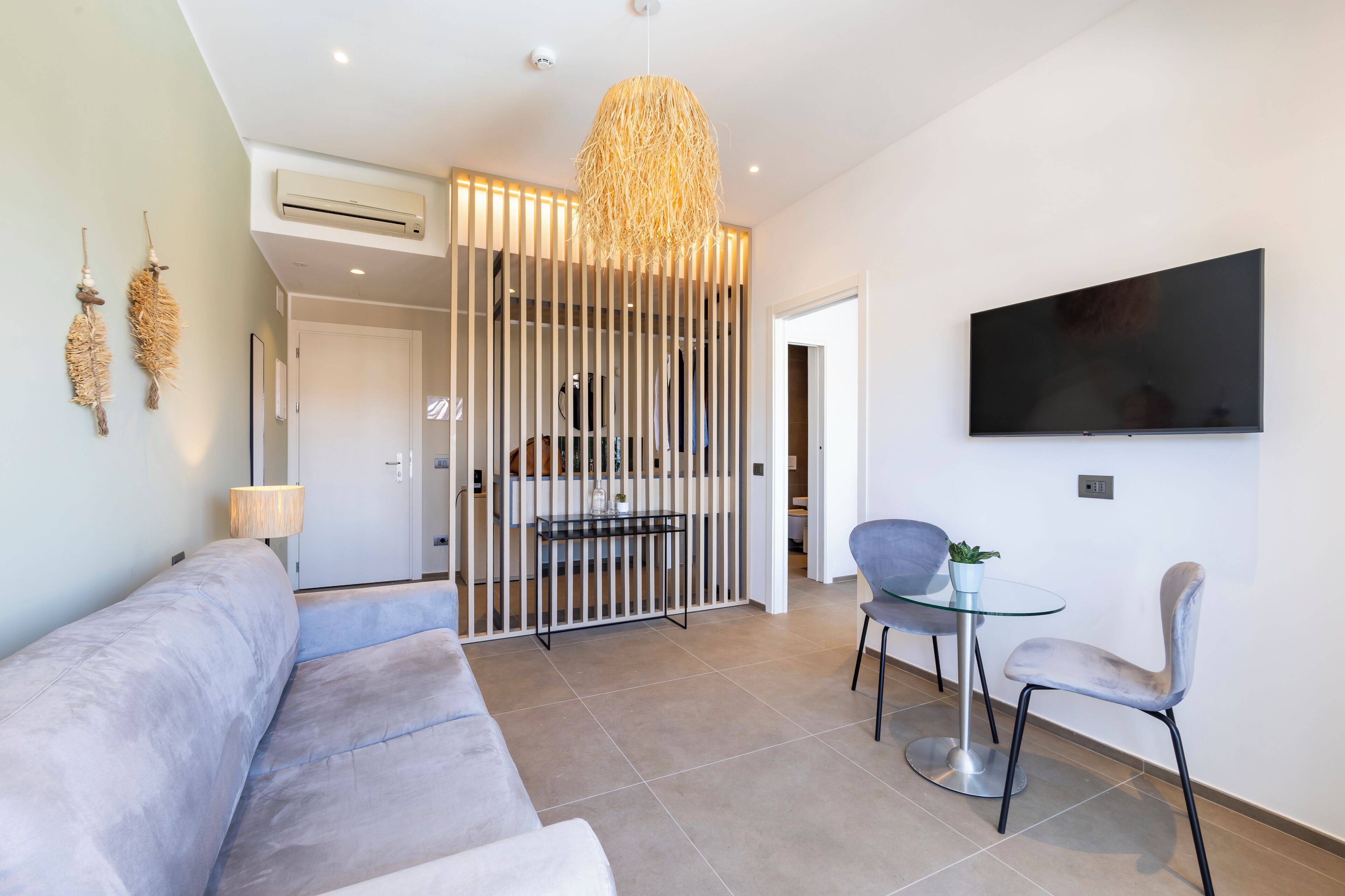Una sala de estar moderna con un sofá, una mesa de vidrio y sillas, un televisor montado en la pared y una lámpara colgante decorativa.