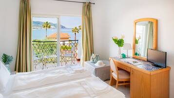 Une chambre d’hôtel comprenant un lit, un bureau avec un ordinateur, une chaise, une petite table et un balcon offrant une vue sur la mer et les palmiers.
