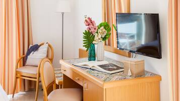 Een hotelkamer met een houten bureau, een stoel, een televisie, een vaas met bloemen en een lamp.