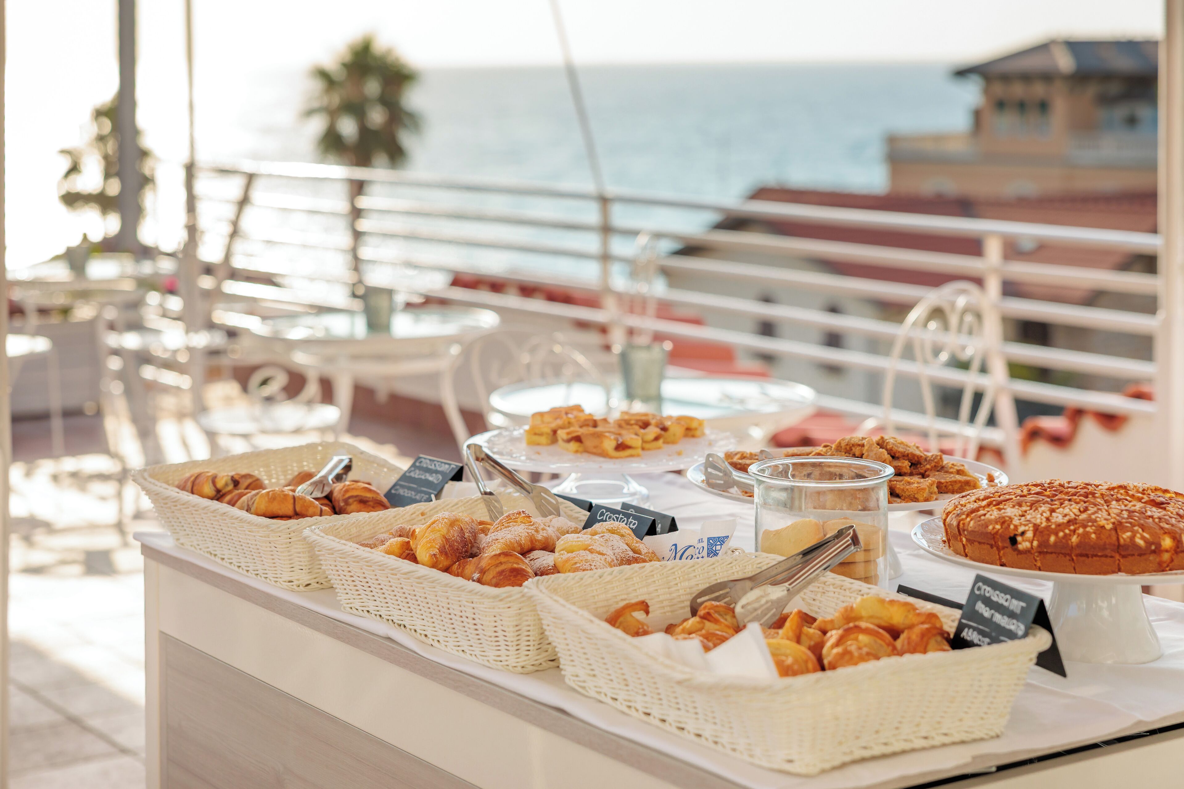 A buffet table with various pastries and bread, set against a backdrop of a balcony with a view of the sea and buildings.
