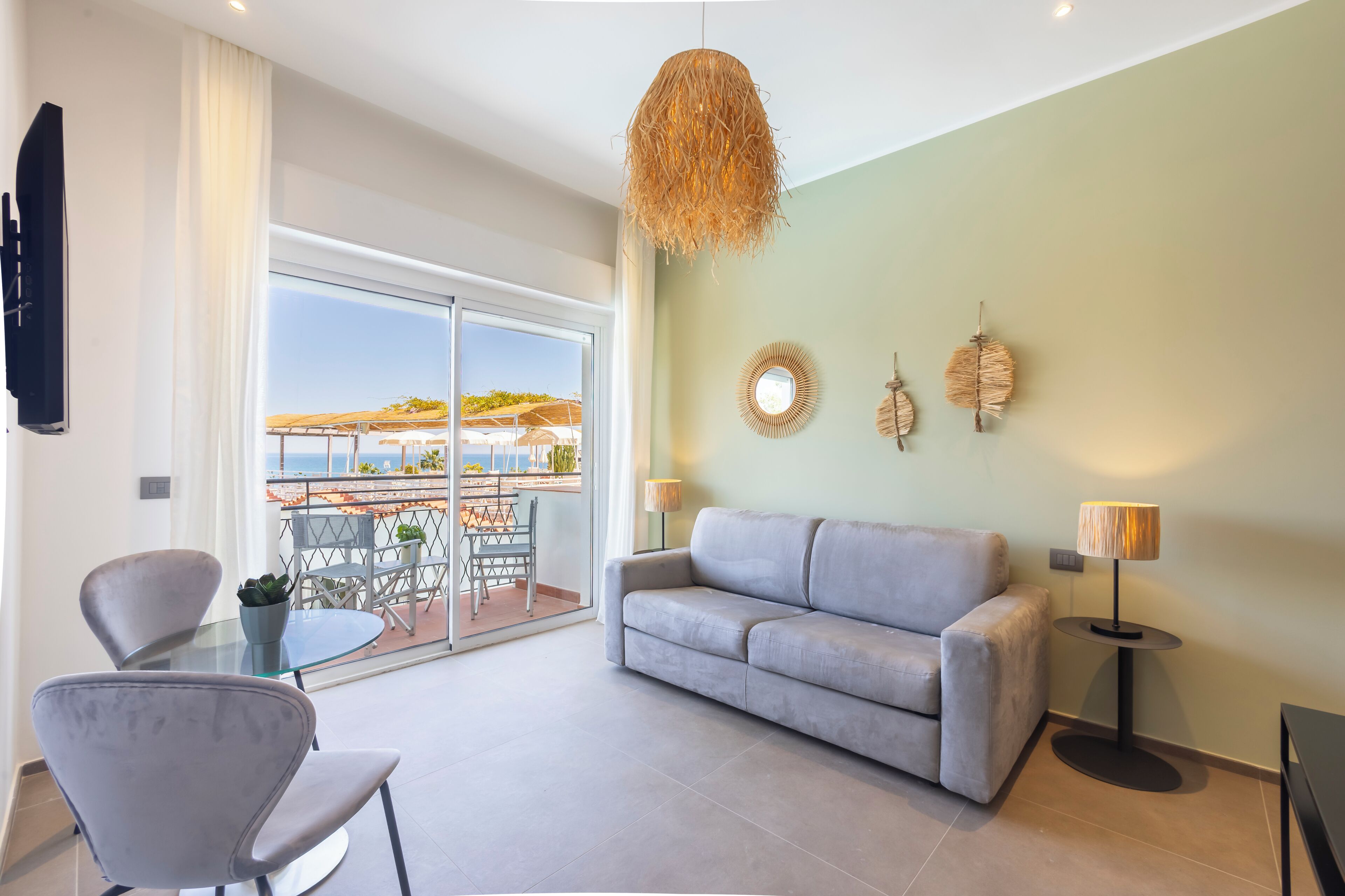 Una sala de estar moderna con un sofá gris, una mesa de vidrio y sillas. Cuenta con un balcón con vista al mar y sombrillas.