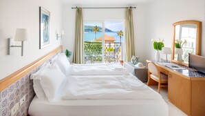 A hotel room with a bed, a desk, a chair, a small table, a TV, and a balcony with a view of the sea.