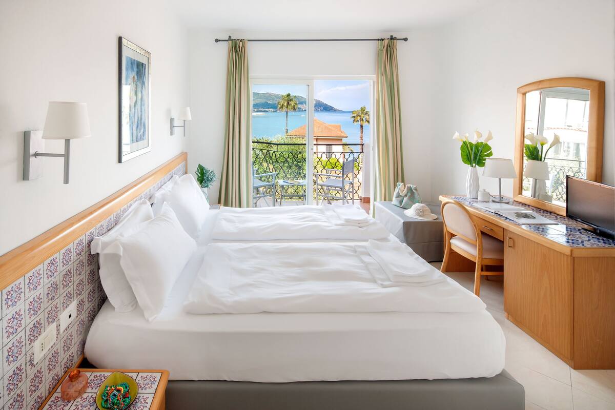 Habitación de hotel con cama, escritorio, silla, mesita, televisor y balcón con vista al mar.