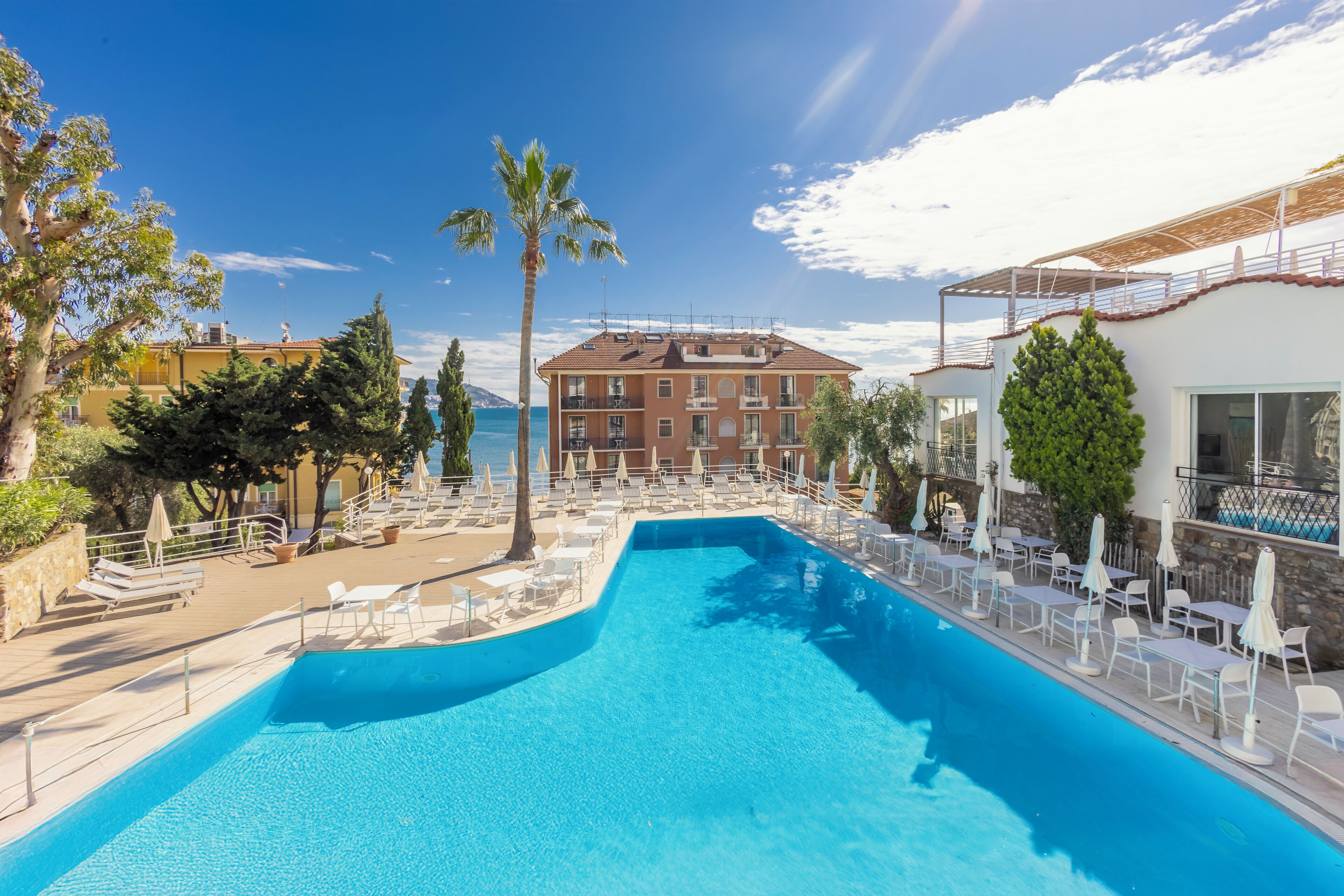 Un espace piscine avec des chaises longues et des parasols, un bâtiment doté de balcons et un ciel d’un bleu éclatant.