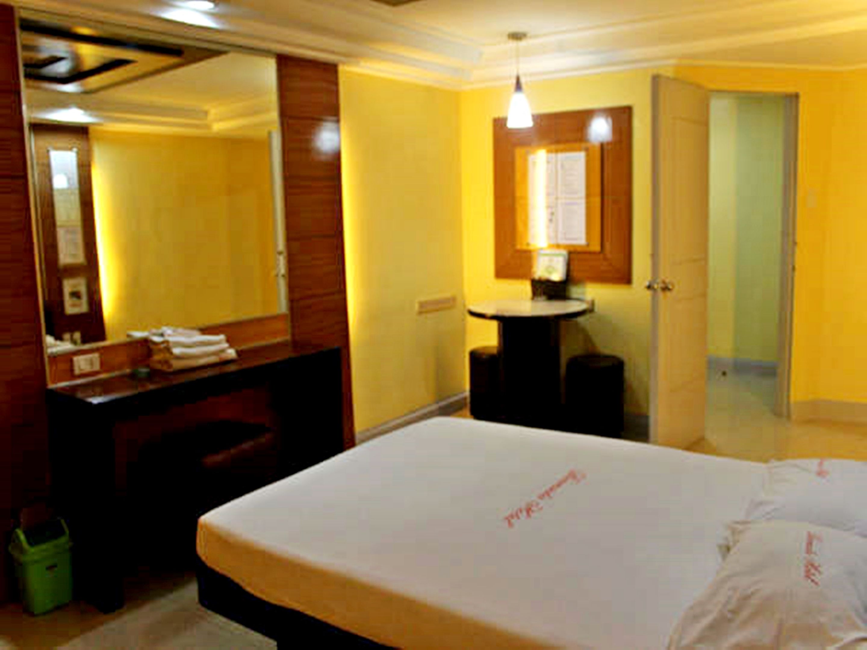 Habitación de hotel con una cama grande, un aparador de madera, un espejo, una mesita con una lámpara y una puerta que da a otra habitación.