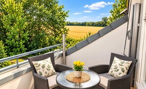 A balcony with wicker furniture and a table, overlooking a field and trees.