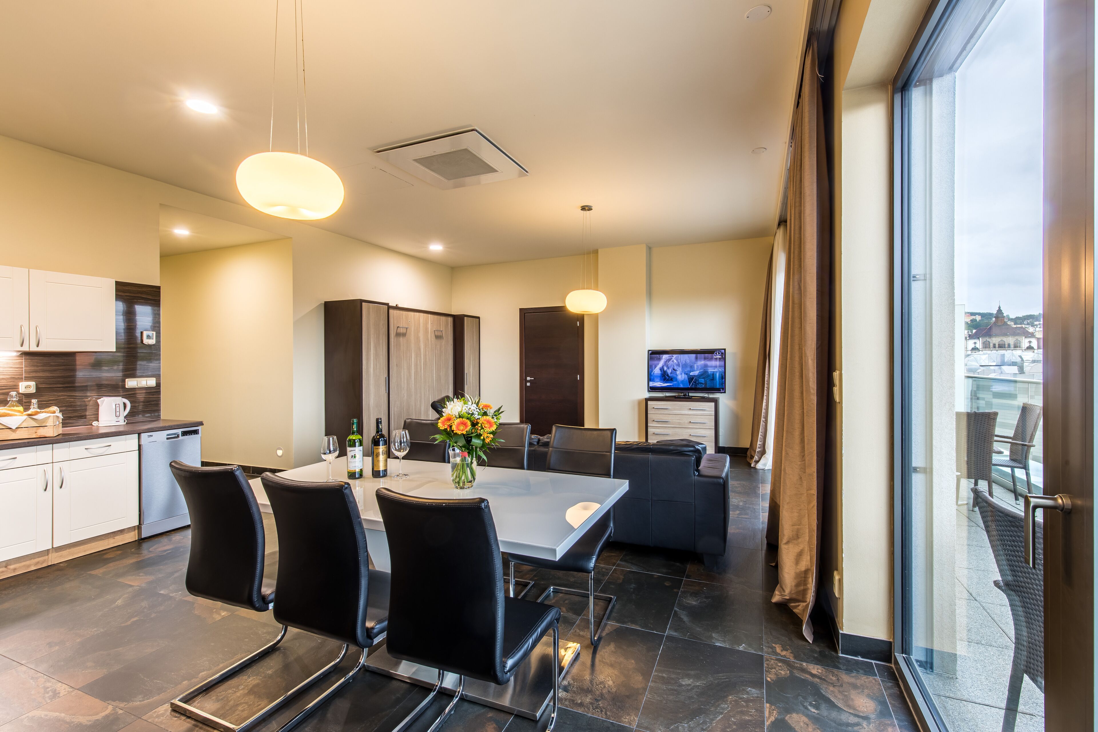 A modern hotel room with a dining area, kitchen, and a large window offering a city view.