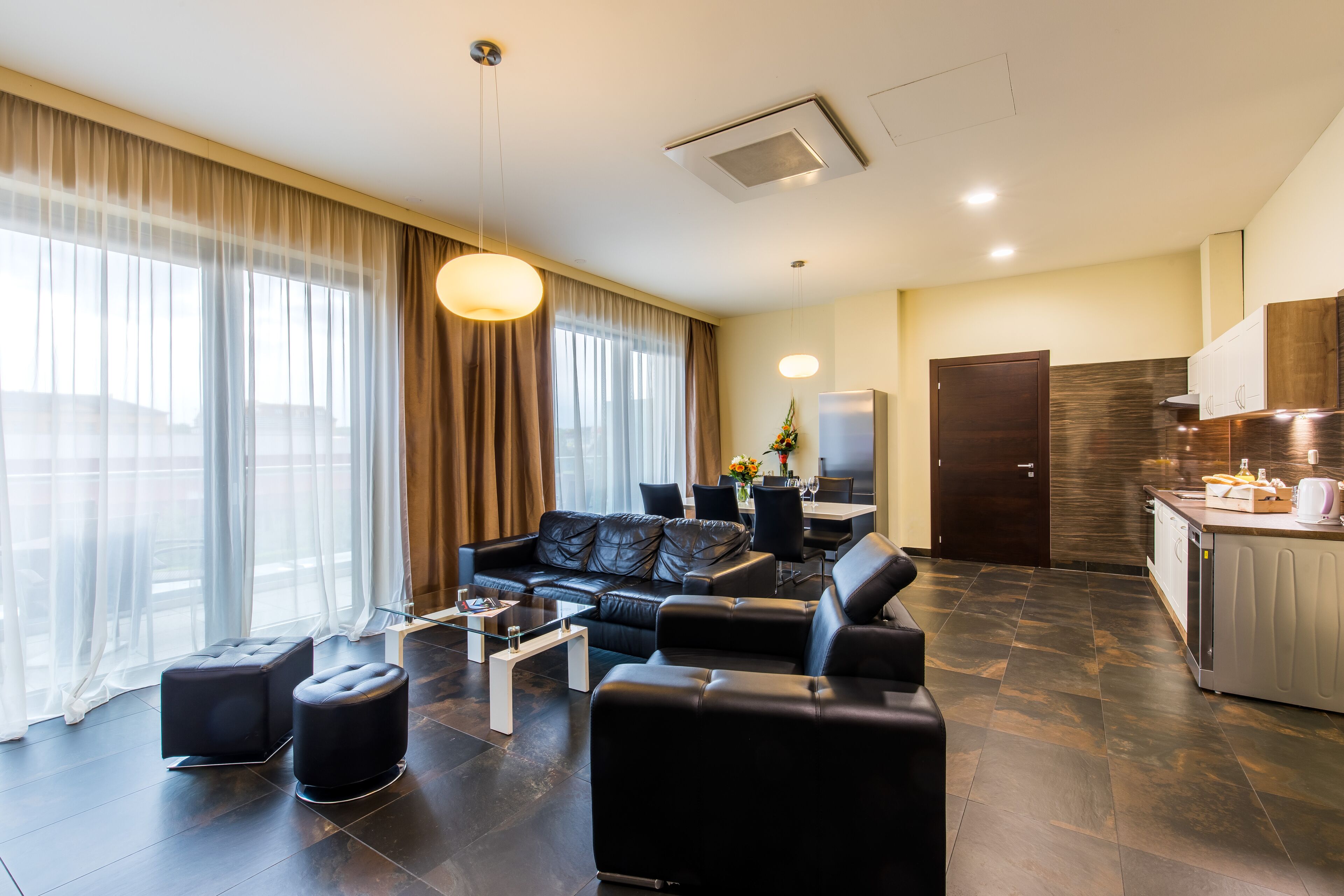 Ein modernes Hotelzimmer mit Wohnbereich, ausgestattet mit einem schwarzen Ledersofa, einem Couchtisch und einem Esstisch mit Stühlen. Es gibt eine Küchenzeile mit Arbeitsplatte und Schränken.