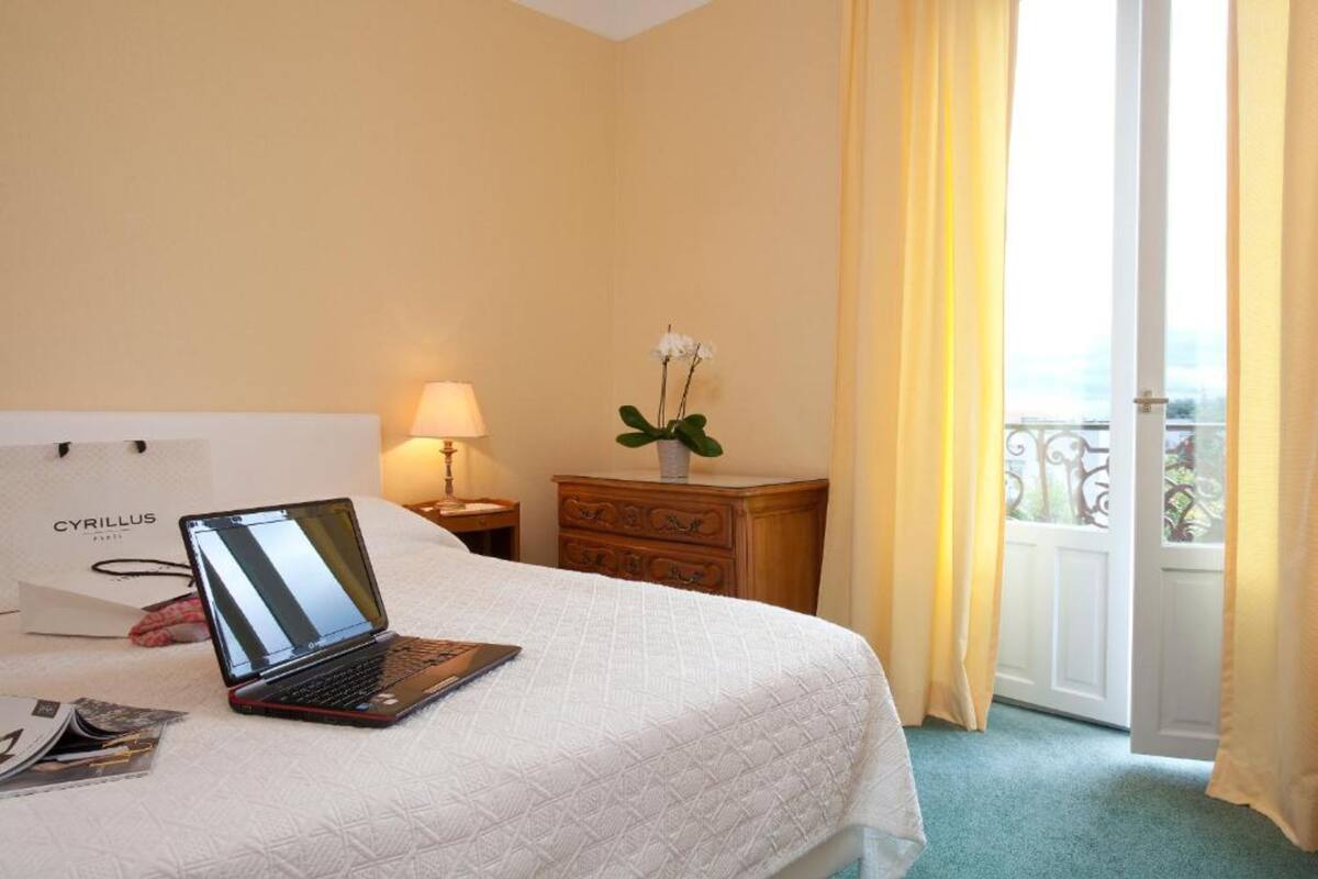 Habitación de hotel con una cama, una laptop, una mesita de noche, un aparador y una ventana con cortinas.