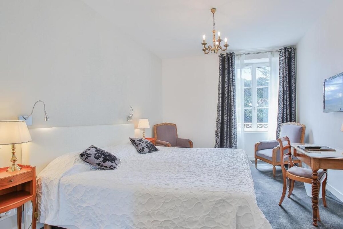 Une chambre d’hôtel avec un grand lit, deux fauteuils, un bureau, un lustre et une fenêtre avec des rideaux.