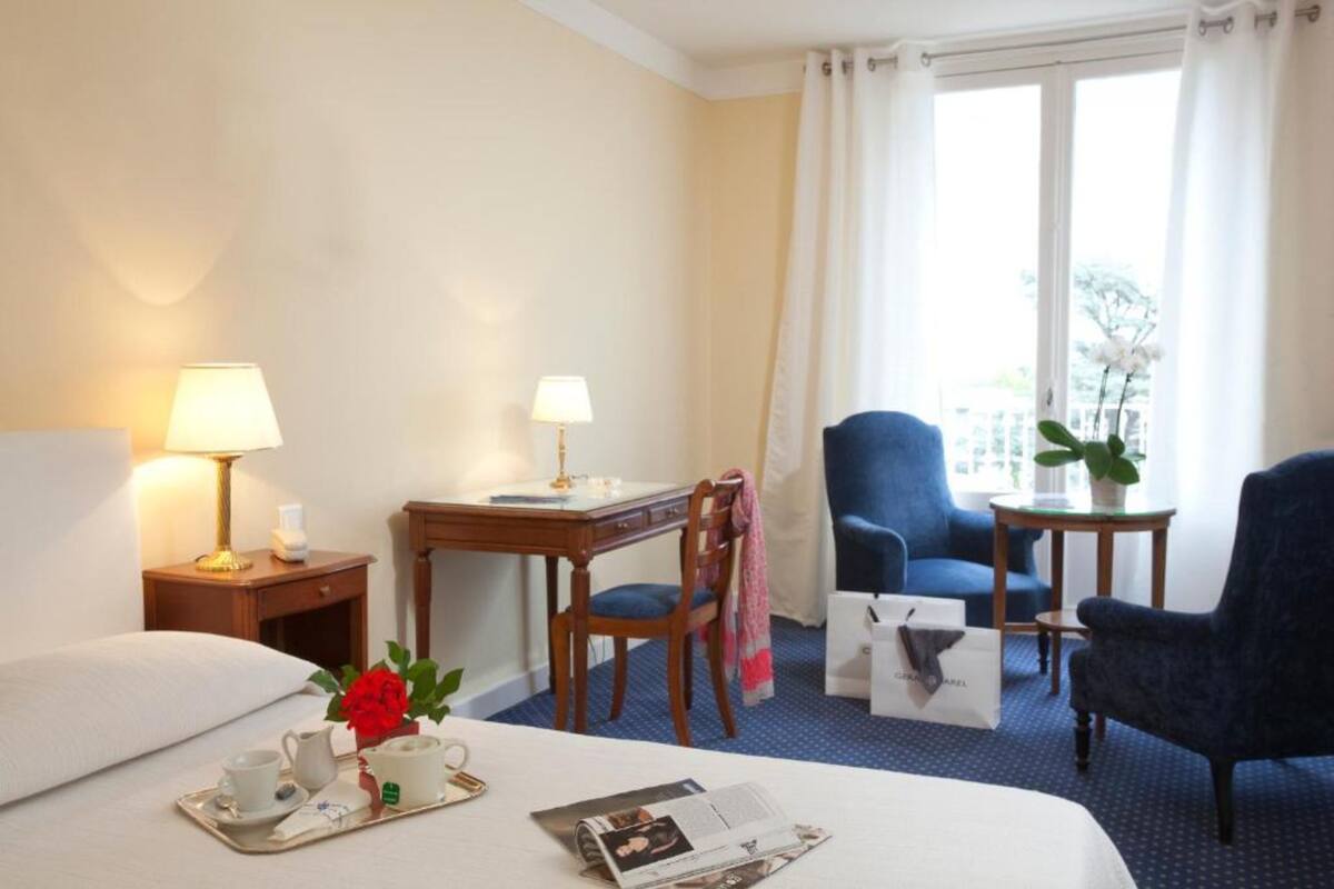 Habitación de hotel con cama, escritorio, dos sillas, una mesita con planta y ventana con cortinas.