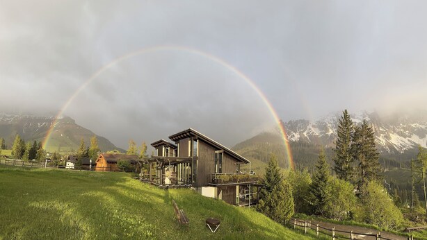 Une maison moderne dotée d’une grande terrasse, entourée d’un paysage verdoyant et de montagnes.