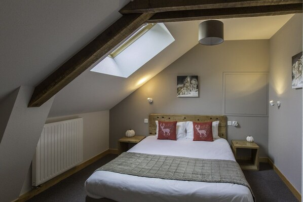 A bedroom with a sloped ceiling, a skylight, a bed with white bedding and decorative pillows, a wooden headboard, bedside tables with lamps, and a radiator. - Les Granges de Bigose (Rimeize)