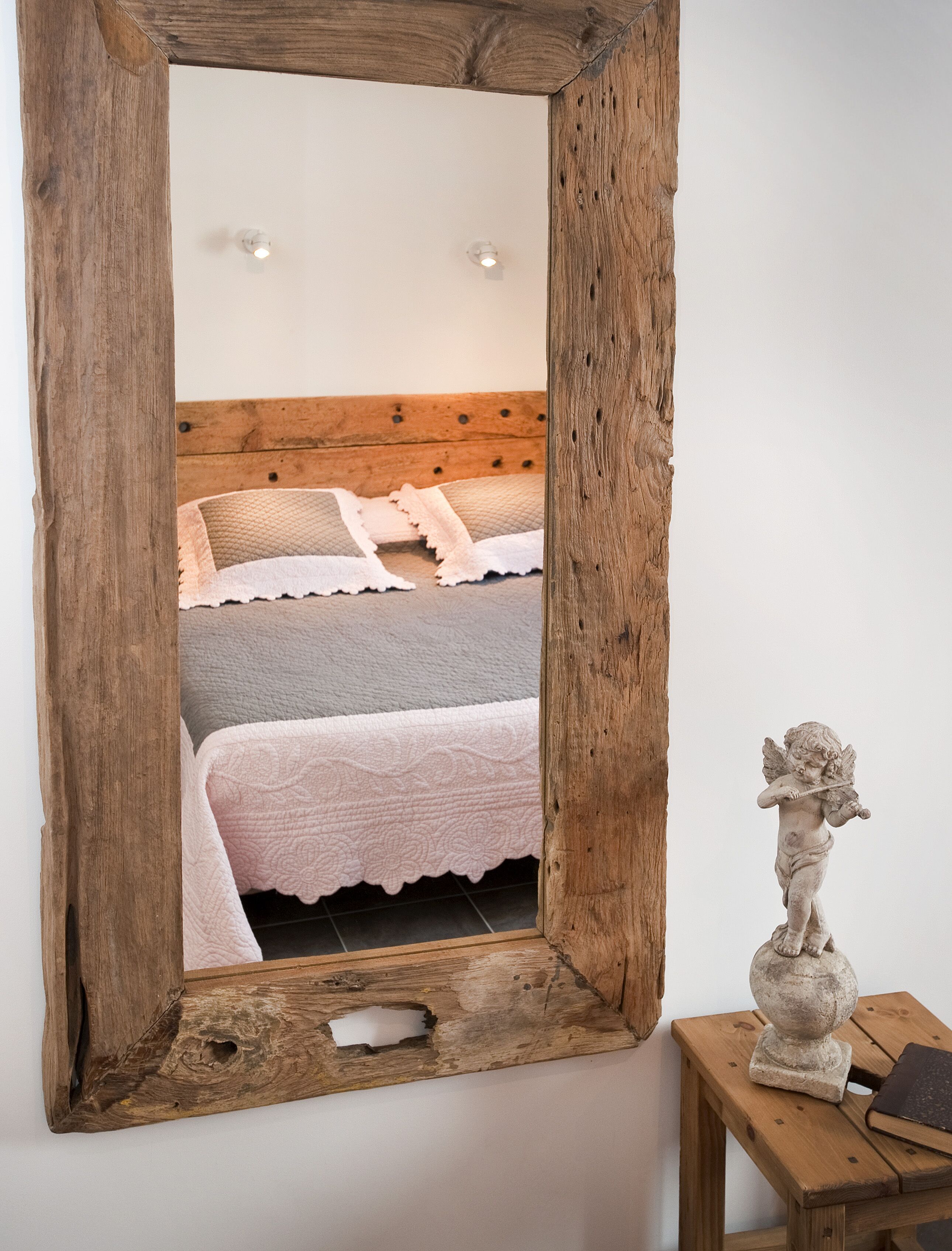 Un espejo con marco de madera rústica refleja un dormitorio con cama, almohadas y una mesita de noche con una estatua.
