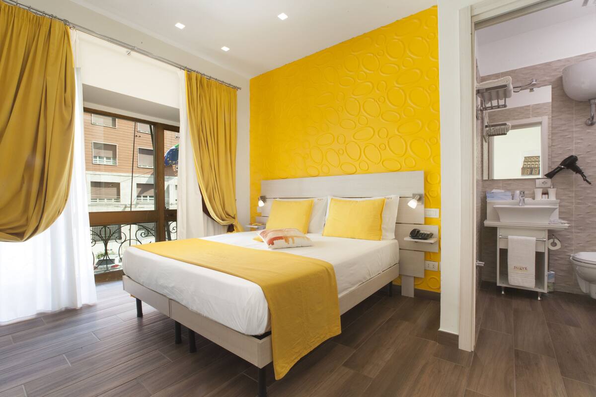 A modern hotel room with a large bed, a bright yellow accent wall, a bathroom with a white sink and mirror, and a balcony with yellow curtains.