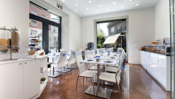 A modern dining area with white furniture, a long table, and a kitchen area with white cabinets and a large window. - Suite Dei Catalani (Naples)