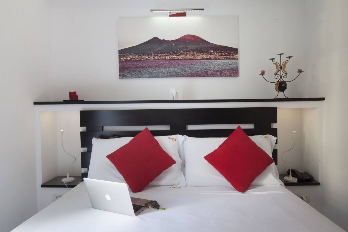 A hotel room with a bed, a laptop, a nightstand with a lamp, a framed picture of a mountain, and a candelabra.